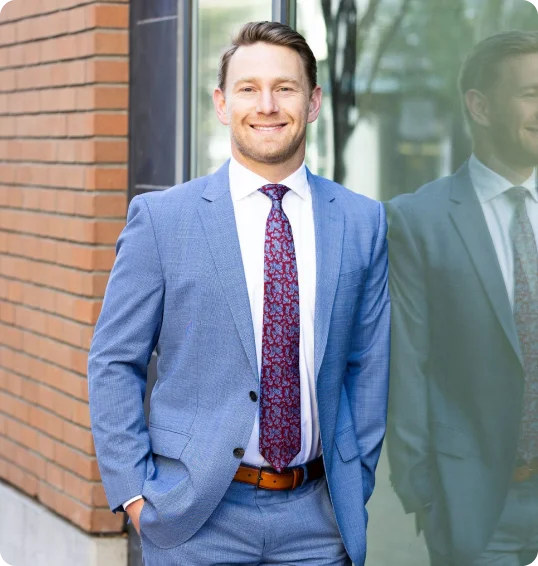 Man in a light blue suit and patterned tie stands outdoors, smiling, with one hand in his pocket next to a glass wall reflecting his image.
